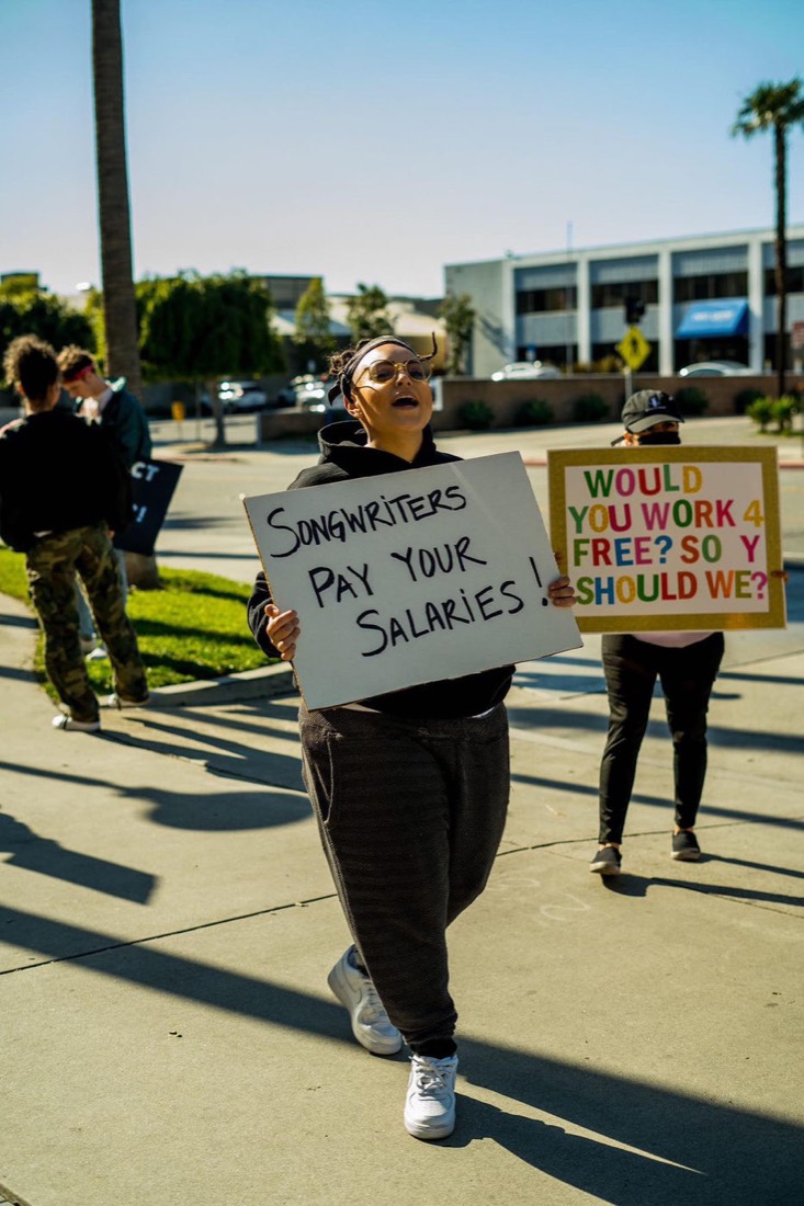 A creator holding a sign that reads “Songwriters pay your salaries”